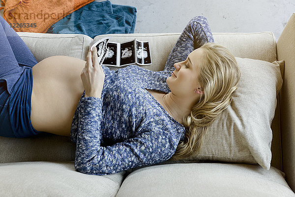 Lächelnde schwangere Frau auf der Couch liegend mit Blick auf Ultraschallbilder