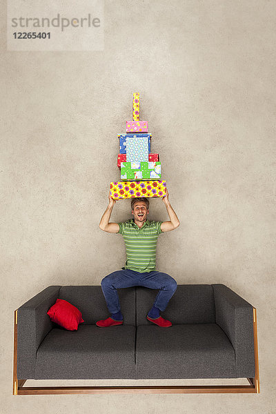 Mann sitzt auf der Couch und balanciert einen Haufen Geschenke auf dem Kopf.
