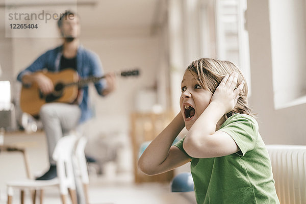 Erschrockener Sohn  der sich die Ohren zuhielt und zu Hause Gitarre spielt.