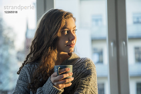 Porträt eines jungen Mädchens mit Kaffeetasse  die abends aus dem Fenster schaut.