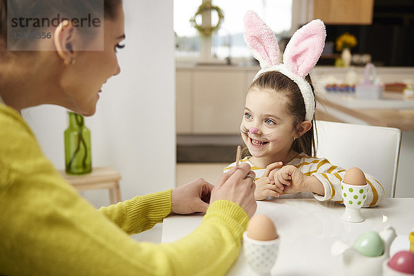 Mädchen mit Hasenohren und Mutter am Tisch sitzend mit Ostereiern