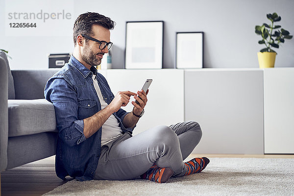 Lächelnder Mann auf dem Boden im Wohnzimmer sitzend mit dem Handy