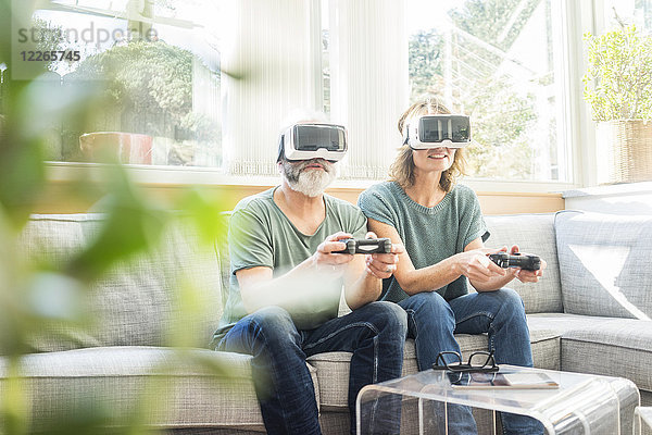 Ein reifes Paar  das zu Hause auf der Couch sitzt und eine VR-Brille trägt und ein Videospiel spielt.