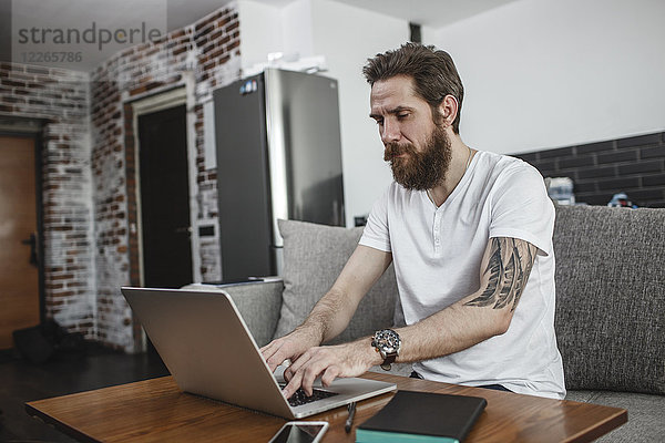 Der bärtige Mann sitzt zu Hause auf der Couch und benutzt einen Laptop.