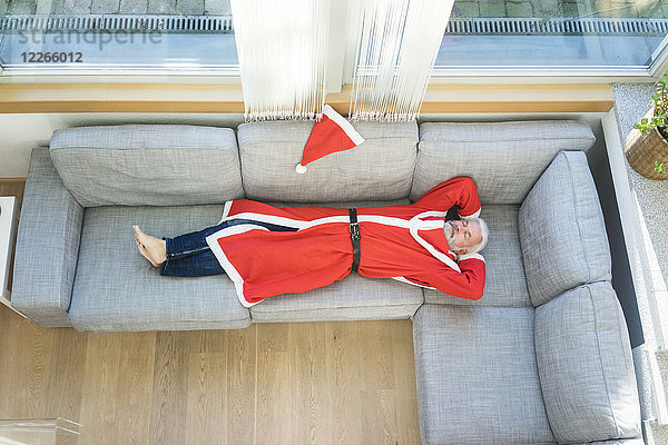 Bärtiger reifer Mann im Weihnachtsmann-Kostüm auf der Couch im Wohnzimmer liegend