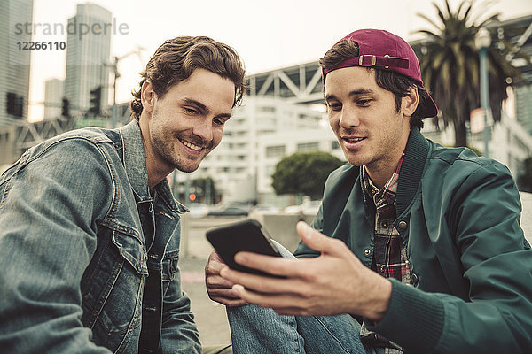 Zwei lächelnde junge Männer teilen sich ihr Handy im Freien.
