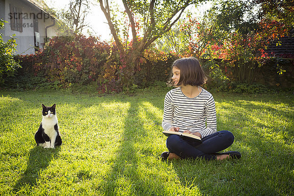 Lächelndes Mädchen sitzt auf der Wiese mit einem Buch neben der Katze.
