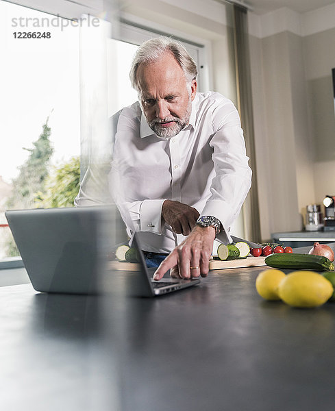 Der reife Mann bereitet das Essen in der Küche zu  während er den Laptop benutzt.