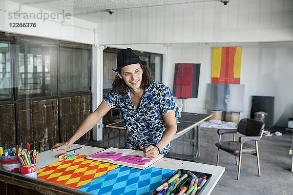Lächelnder Künstler bei der Arbeit  Zeichnen in einem Notizbuch in seinem Loft-Atelier mit Bildern und Kunstwerken im Hintergrund.