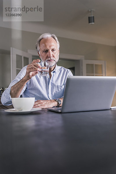 Nachdenklicher reifer Mann sitzt am Tisch mit einem Glas Wasser und schaut auf den Laptop.
