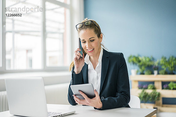 Geschäftsfrau sitzt am Schreibtisch  spricht am Telefon  schaut auf das digitale Tablett