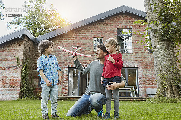 Vater mit zwei Kindern  die mit einem Spielzeugflugzeug im Garten ihres Hauses spielen.