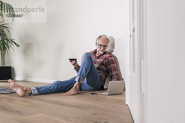 Barfußmann sitzt zu Hause auf dem Boden und entspannt sich mit Laptop und einem Glas Rotwein.