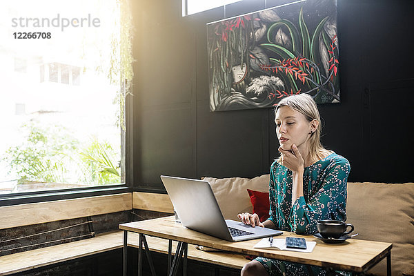 Junge Frau mit grünem Kleid sitzt im Café und arbeitet an ihrem Laptop.