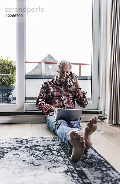 Barfuss-Mann am Telefon entspannt auf dem Boden zu Hause mit Laptop und einem Glas Rotwein
