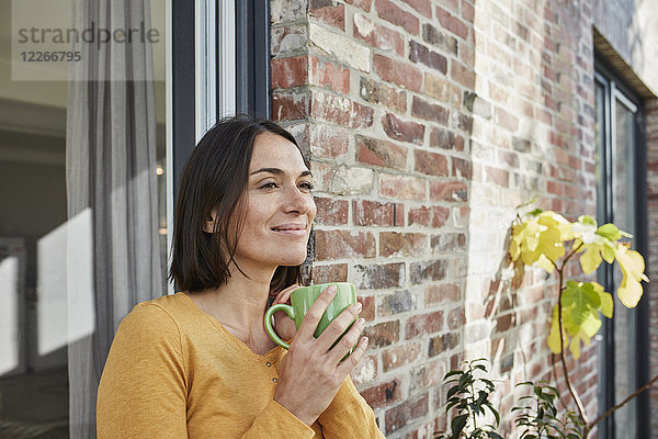 Lächelnde Frau trinkt aus Tasse vor ihrem Haus