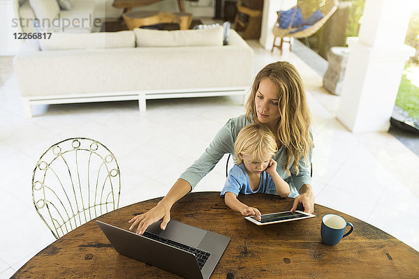 Junge sitzt auf dem Schoß seiner Mutter und schaut auf eine Tafel  während seine Mutter an einem Laptop arbeitet.