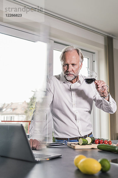 Portrait eines reifen Mannes mit einem Glas Rotwein in der Küche mit Blick auf den Laptop