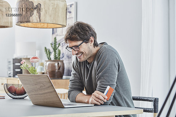 Lächelnder Mann hält eine Karte mit einem Laptop auf dem Tisch zu Hause.