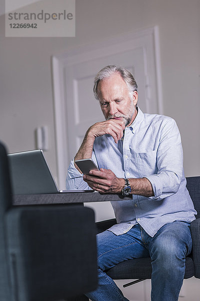 Erwachsener Mann am Tisch sitzend mit Laptop beim Blick aufs Handy