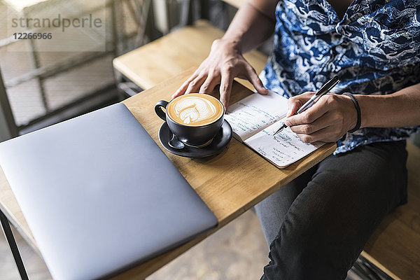 Künstlerin schreibt in ein Notebook in einem Café mit Kaffee und Laptop auf dem Tisch