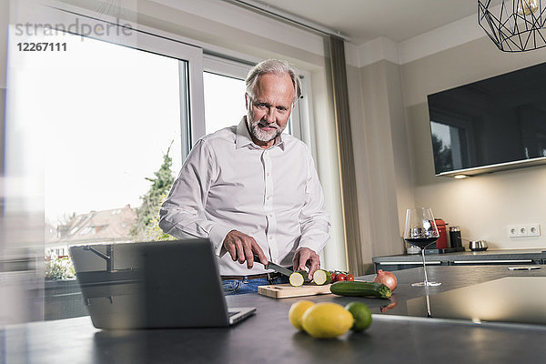 Der reife Mann bereitet das Essen in der Küche zu  während er sich den Laptop ansieht.