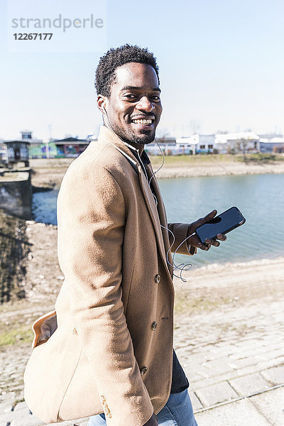 Porträt des lächelnden Geschäftsmannes am Wasser mit Handy und Kopfhörer