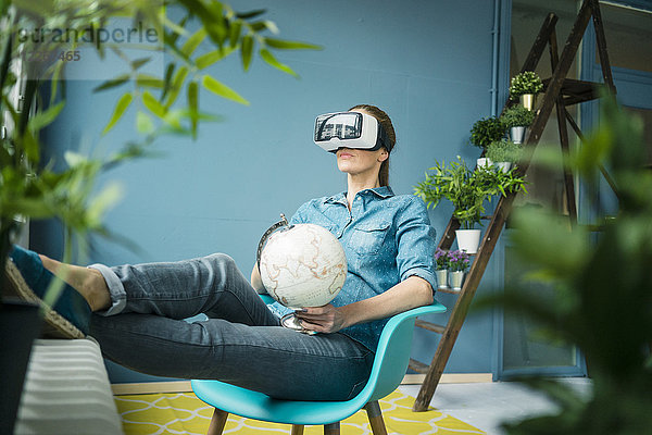 Schöne Frau in ihrem Haus sitzend  mit Pflanzen geschmückt  Globus haltend  VR-Brille tragend