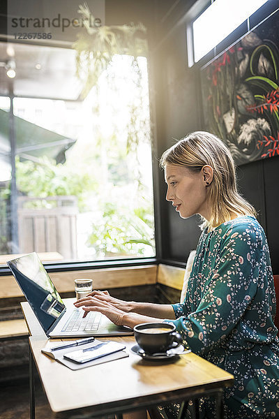 Junge Frau mit grünem Kleid sitzt im Café und arbeitet an ihrem Laptop.