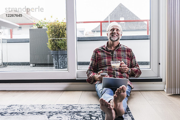 Fröhlicher Mann  der sich zu Hause mit Laptop  Handy und einer Tasse weißem Kaffee auf dem Boden entspannt