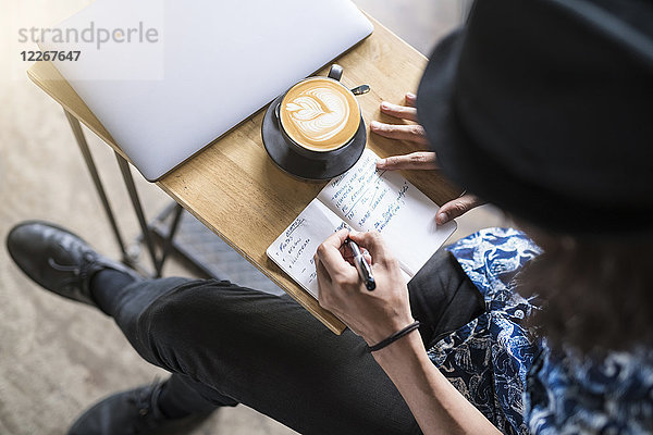 Künstlerin schreibt in ein Notebook in einem Café mit Kaffee und Laptop auf dem Tisch
