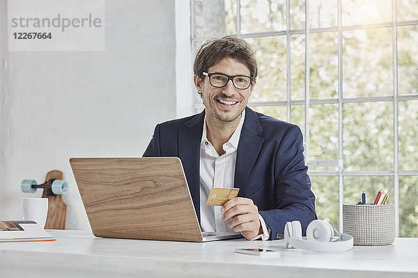 Porträt des lächelnden Geschäftsmannes mit Laptop auf dem Schreibtisch mit Kreditkarte