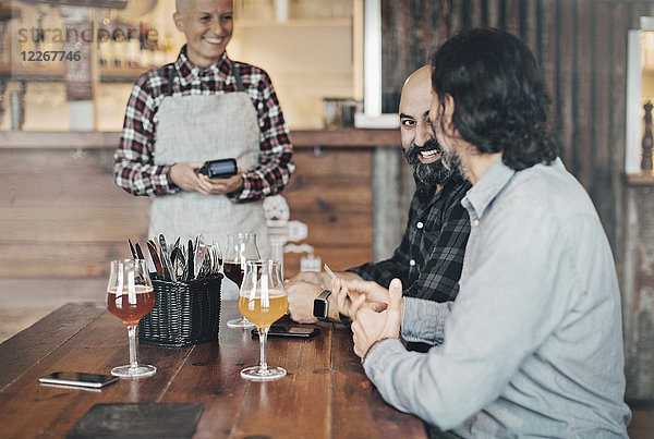 Barkeeperin mit Kreditkartenleser im Gespräch mit männlichen Kunden am Tisch in der Bar
