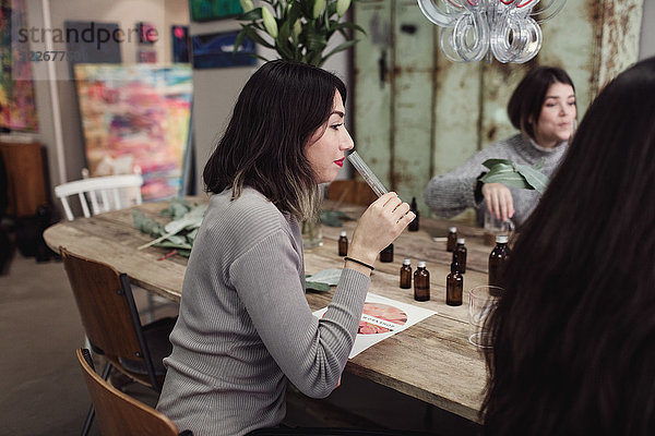 Seitenansicht einer jungen Frau  die beim Sitzen mit Kollegen am Tisch in der Werkstatt Flüssigkeit aus dem Reagenzglas riecht.