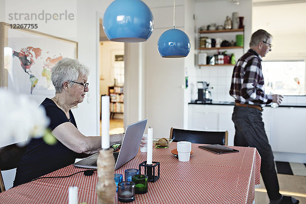 Seitenansicht der älteren Frau  die mit Laptop am Esstisch sitzt  während der Mann im Hintergrund läuft.