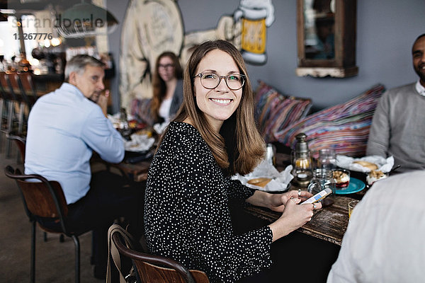 Porträt einer lächelnden Frau  die ein Handy hält  während sie mit Freunden am Tisch sitzt.