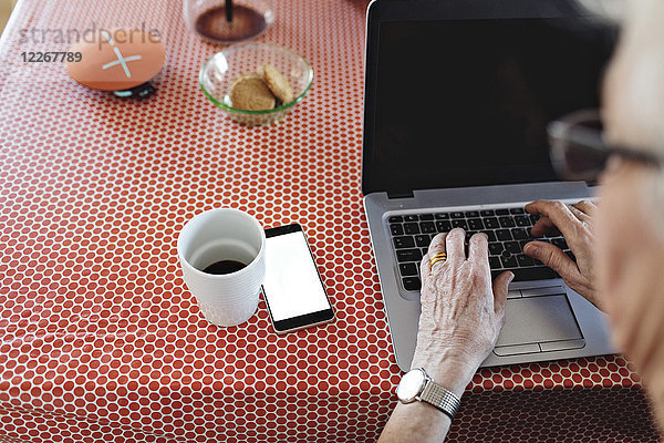 High-Winkel-Ansicht der Seniorin mit Laptop per Smartphone und Kaffeetasse auf dem Esstisch zu Hause