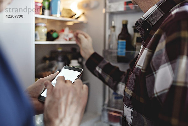 Seniorenfrau mit Smartphone steht in der Küche zu Hause neben dem pensionierten Mann