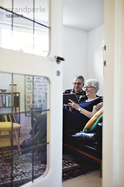 Volle Länge des lächelnden Seniorenpaares auf dem Sofa mit digitalem Tablett von der Tür aus gesehen.