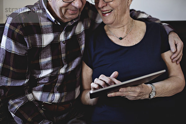 Mittelteil eines lächelnden älteren Paares  das zu Hause sitzt und sich ein digitales Tablett teilt.