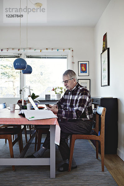 Seitenansicht des pensionierten älteren Mannes  der das Smartphone benutzt  während er mit den Finanzrechnungen am Esstisch sitzt.