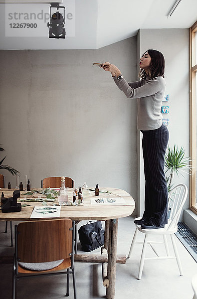 Seitenansicht der jungen Frau  die auf einem Stuhl steht und Parfümflaschen über dem Tisch in der Werkstatt fotografiert.