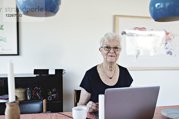 Porträt einer lächelnden Seniorin im Ruhestand mit Laptop am Esstisch