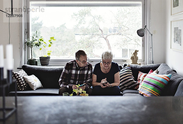 Senioren-Paar mit Smartphones auf dem Sofa im Wohnzimmer gegen das Fenster zu Hause sitzen