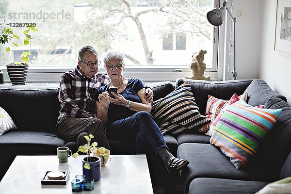 Seniorenpaar teilt sich ein Smartphone auf dem Sofa im Wohnzimmer gegen das Fenster zu Hause.