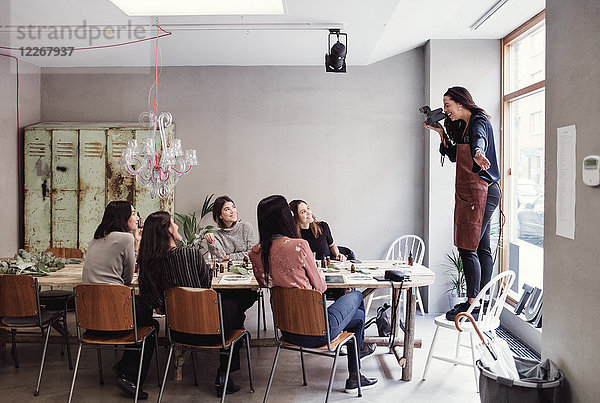 Fröhliche Frau auf dem Stuhl stehend  die weibliche Kollegen am Tisch in der Werkstatt fotografiert.