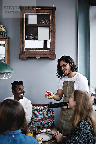 Lächelnder Besitzer serviert Essen für junge Männer und Frauen am Esstisch.