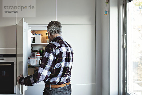 Rückansicht des pensionierten Senioren an der offenen Kühlschranktür in der Wohnküche