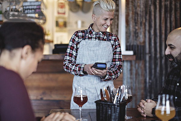 Lächelnde Barkeeperin mit Kreditkartenleser im Gespräch mit Kunden am Tisch in der Bar