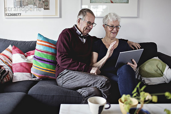 Lächelndes Seniorenpaar sitzt auf dem Sofa und teilt sich ein digitales Tablett im Wohnzimmer zu Hause.